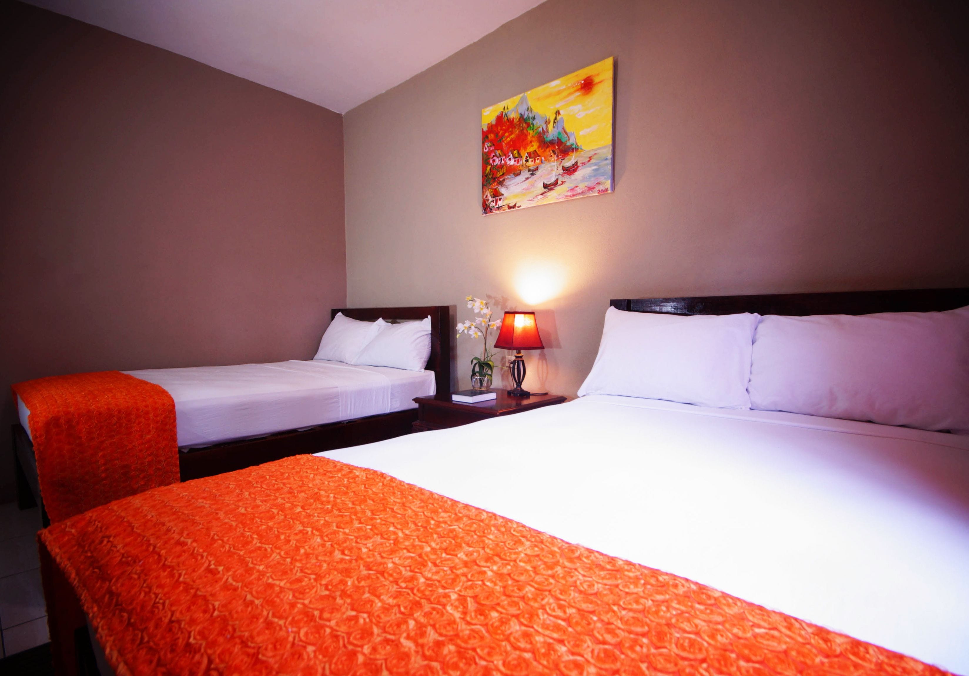 A hotel room with two beds, a painting on the wall, and a nightstand with a lamp and a plant.
