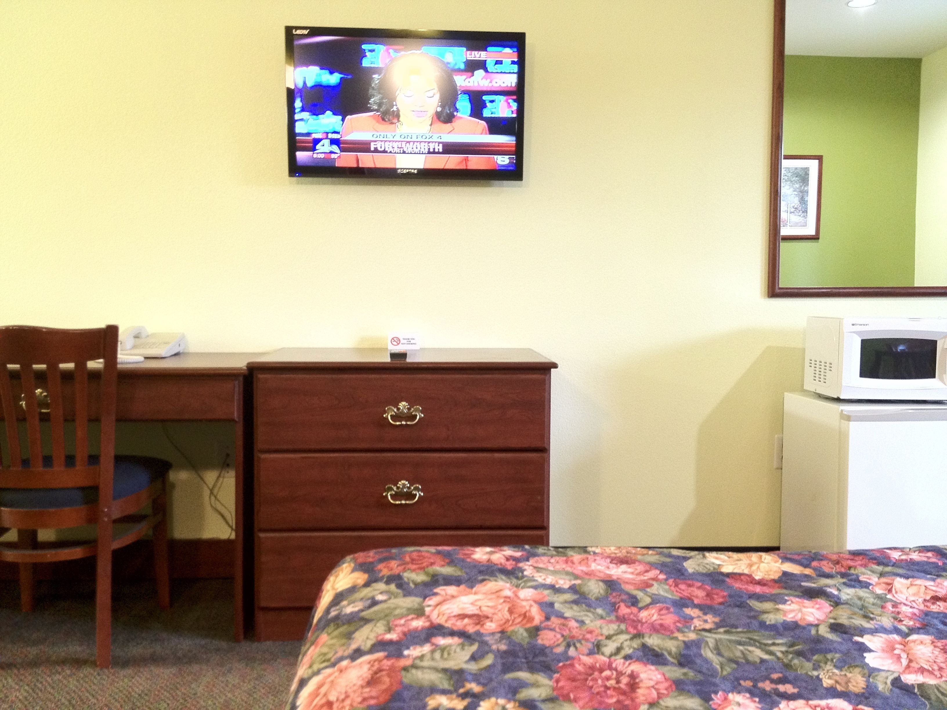 A hotel room with a television mounted on the wall displaying a news program, a wooden desk with a chair, a dresser with drawers, a microwave on a small table, and a floral-patterned bedspread.