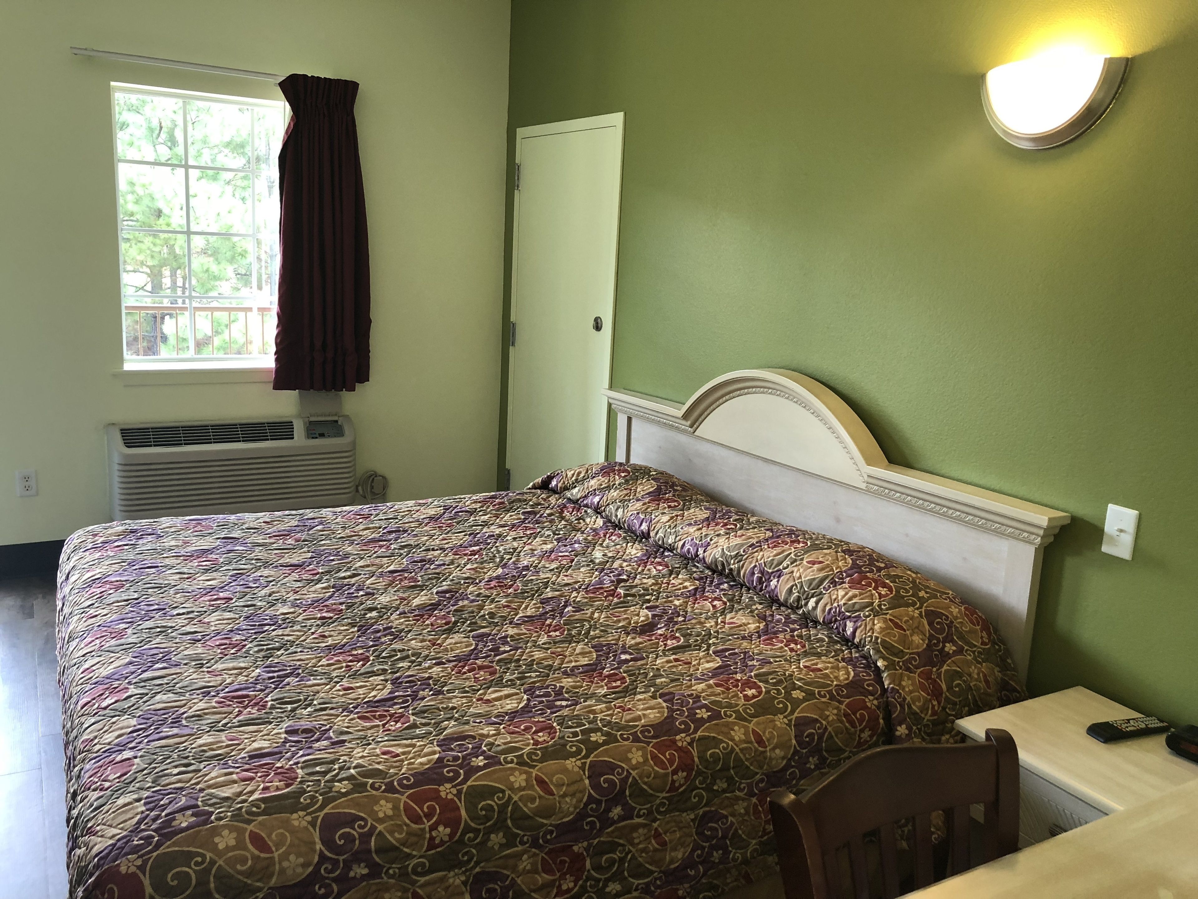 A room with a bed, a table, a chair, a window with curtains, an air conditioning unit, and a wall-mounted light fixture.