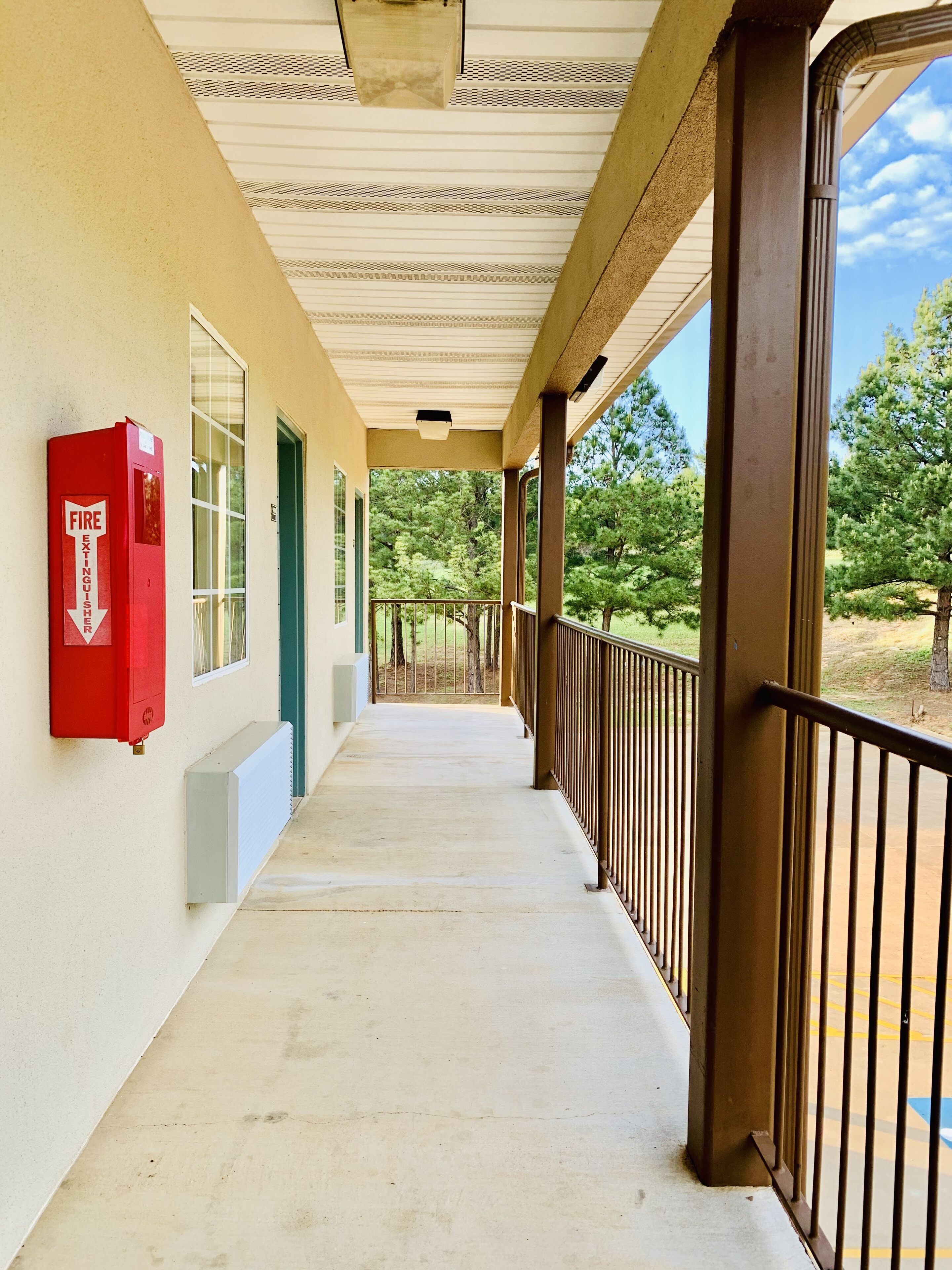 A covered corridor with a fire extinguisher mounted on the wall, a view of trees and greenery outside, and a metal railing along the right side.