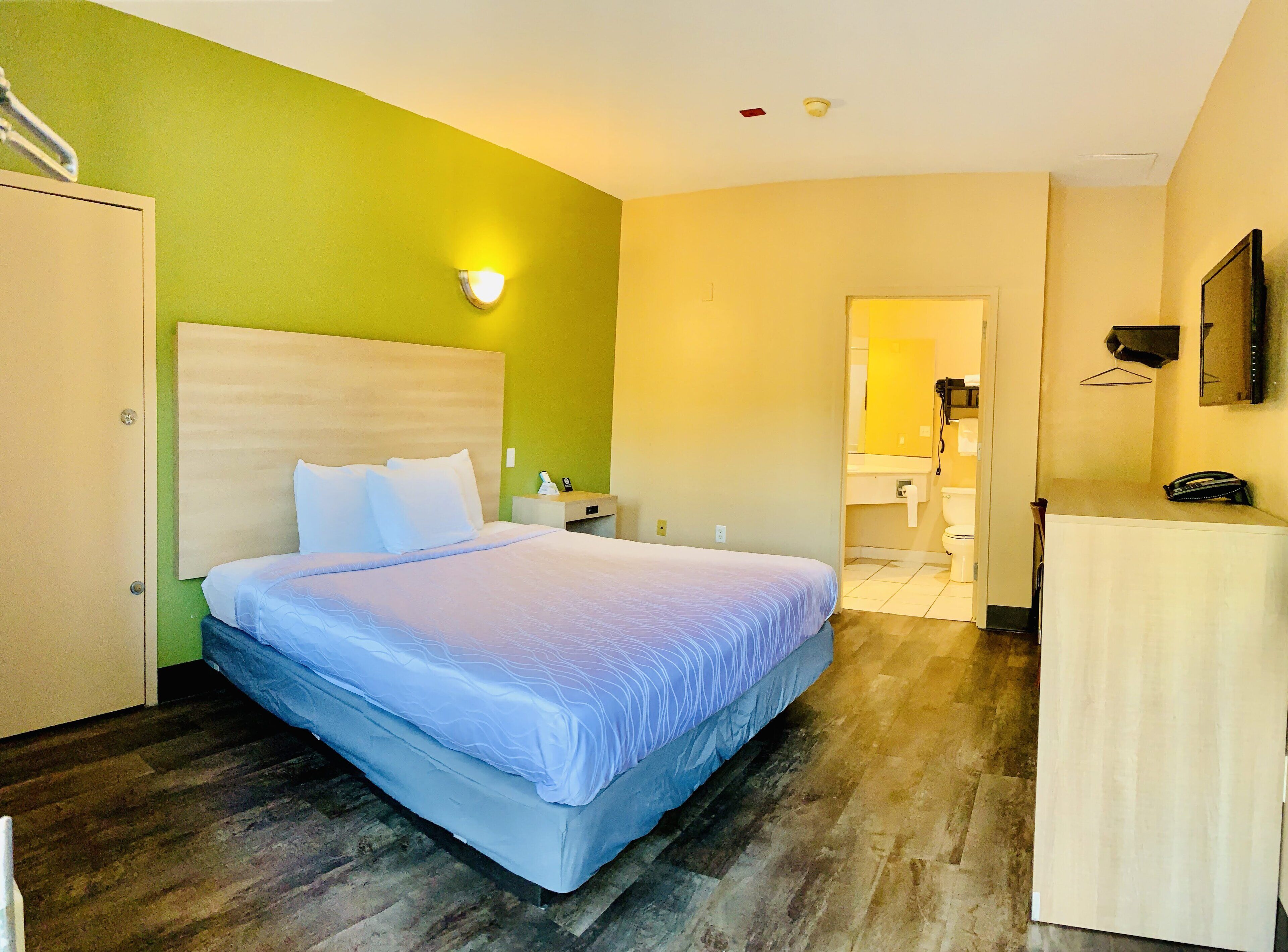 A hotel room with a bed, a nightstand, a desk, a mirror, and a bathroom visible through an open door.