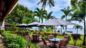An outdoor seating area with tables and chairs, surrounded by lush greenery and palm trees, overlooking a body of water. - Dolphin Beach Bali (Banjar)