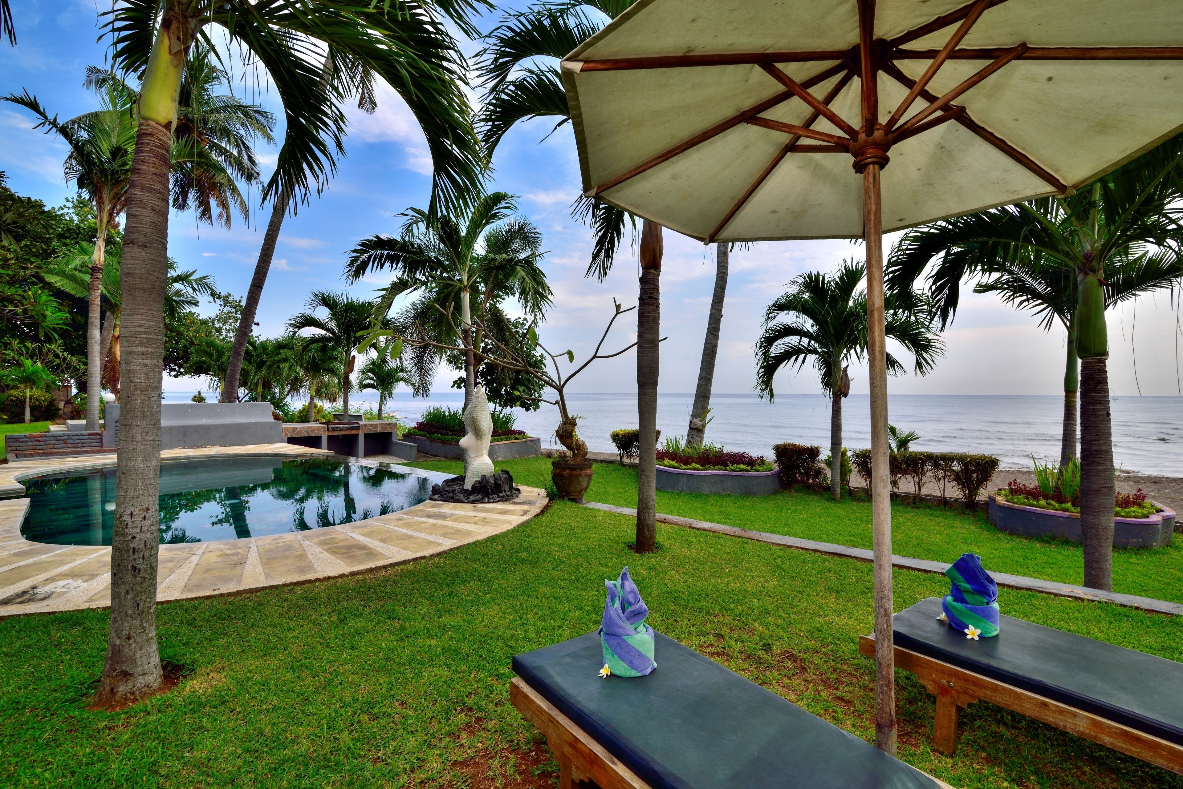 A pool area with a lounge chair and umbrella, surrounded by palm trees and a view of the ocean.