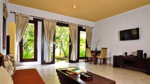 A living room with a sofa, a coffee table with books, a TV, and a view of greenery through the windows. - Dolphin Beach Bali (Banjar)