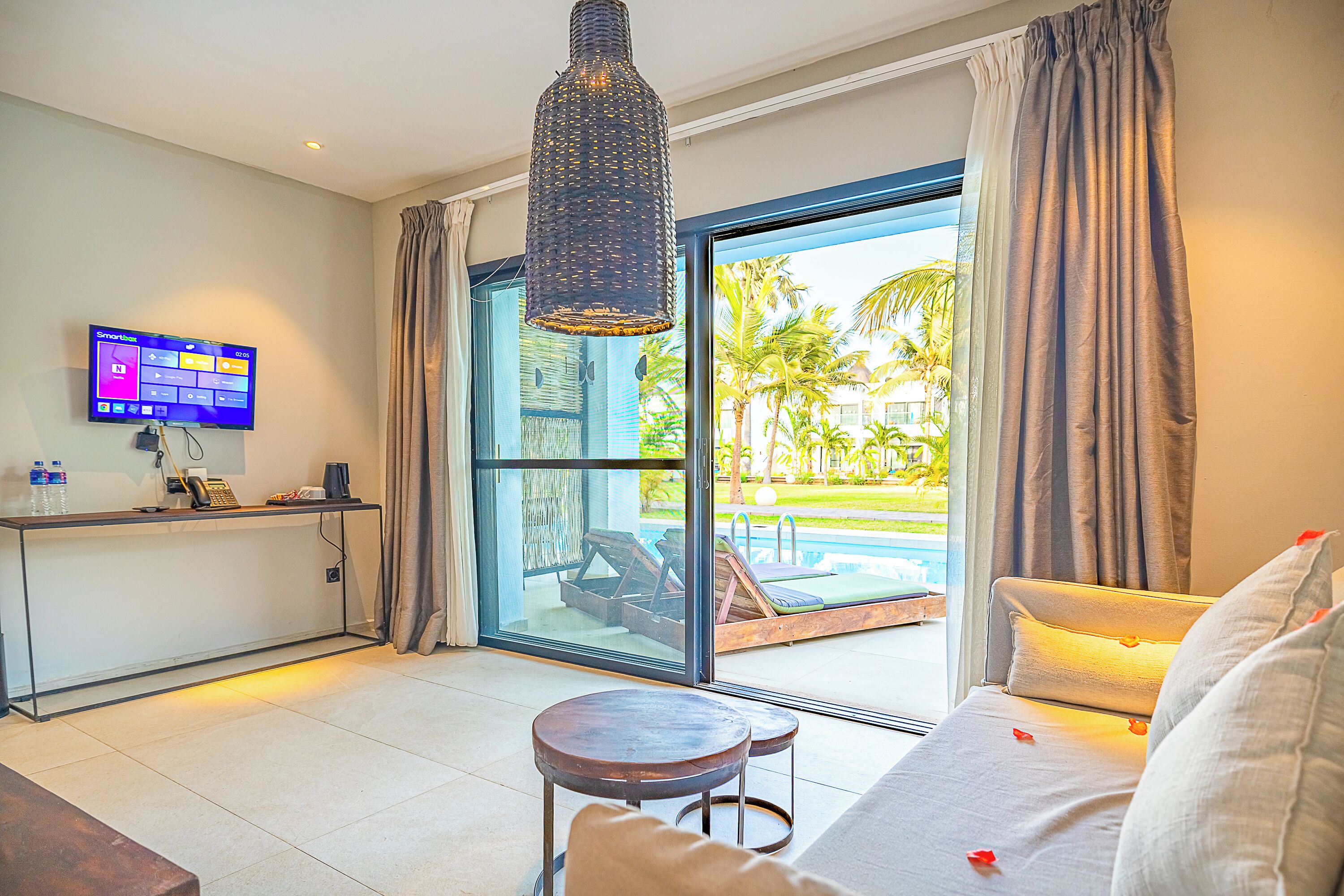 Habitación de hotel moderna con un gran ventanal corredero a una piscina, televisor de pantalla plana, escritorio con teléfono y una zona de estar acogedora con sofá y una mesita redonda.