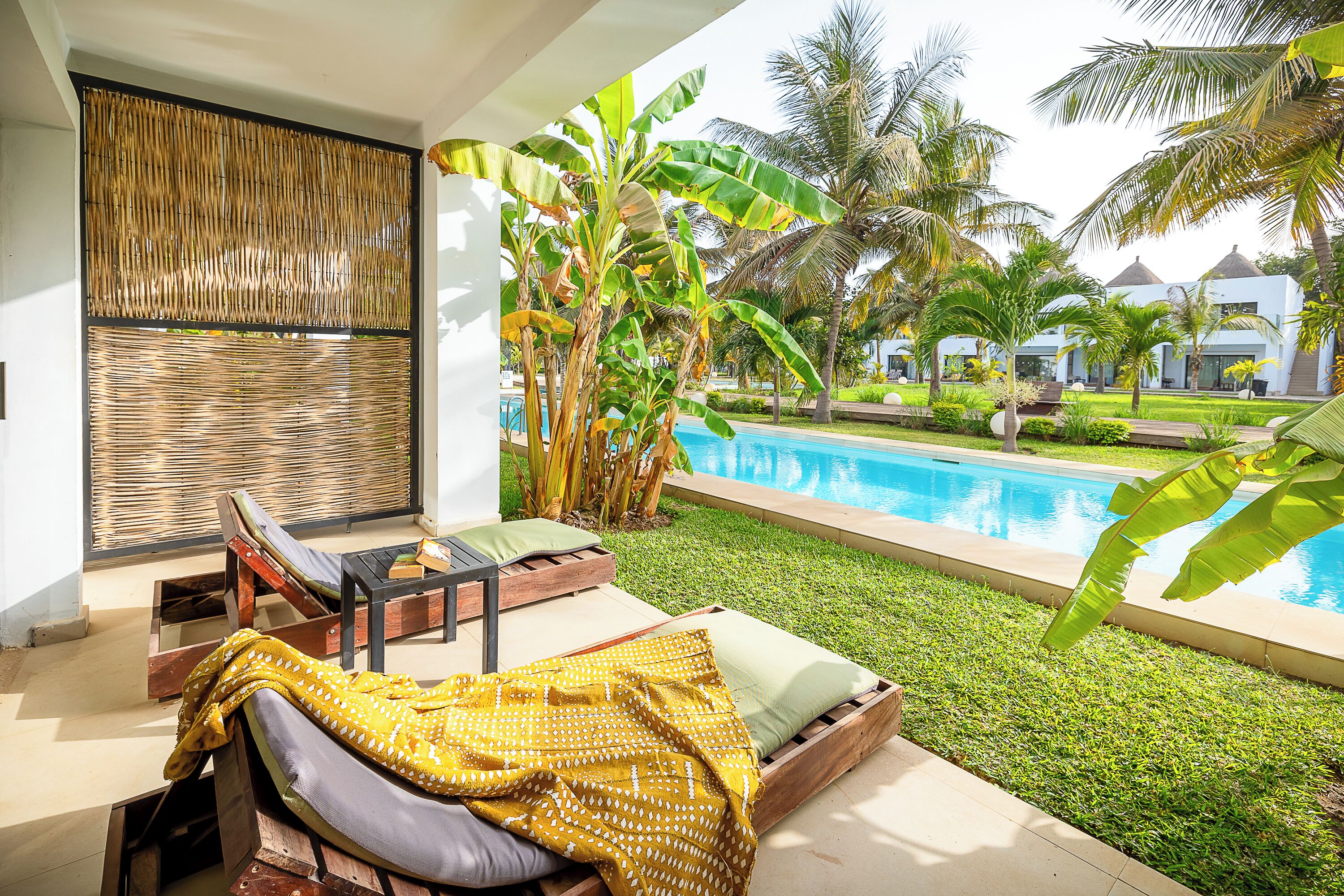 Zona junto a la piscina con una silla de descanso, una mesita y vistas a una piscina rodeada de vegetación y palmeras.