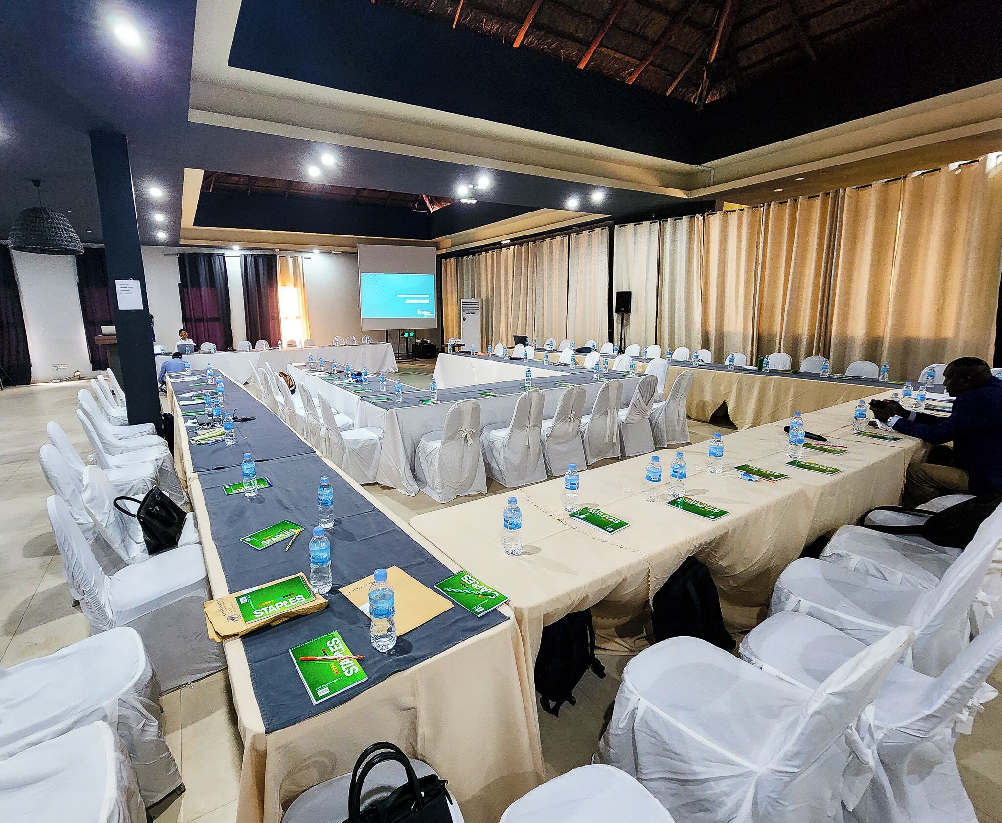 A conference room with long tables, white chairs, and a screen at the front.
