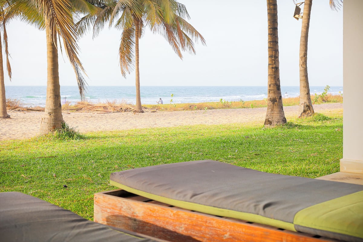 A beach view with palm trees, a sandy beach, and a grassy area with a bed.