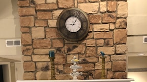 Uma lareira de pedra com um relógio acima, uma peça decorativa sobreposta e uma fogueira acesa.