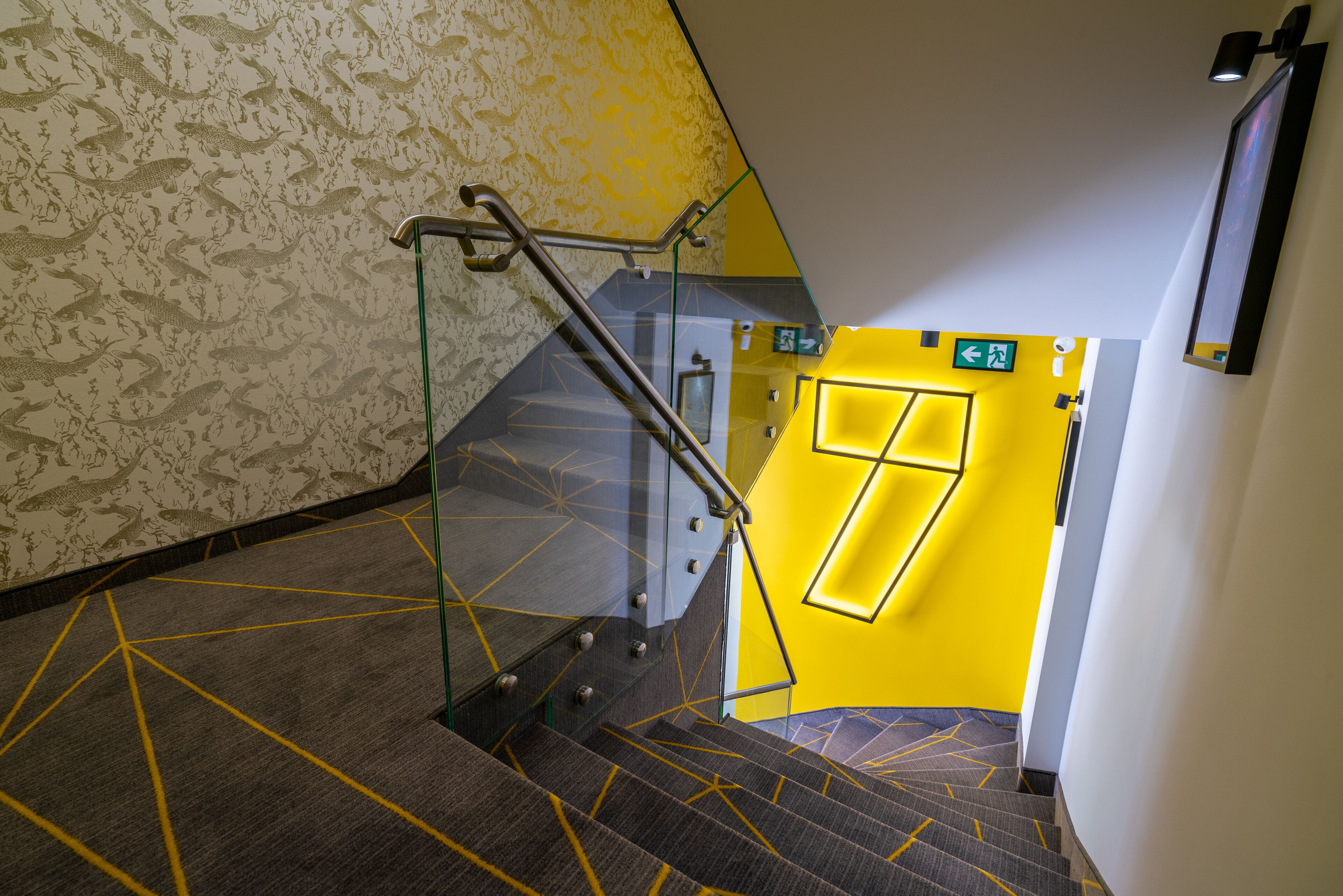 A staircase with geometric carpet patterns, a glass railing, and a yellow door with an exit sign.