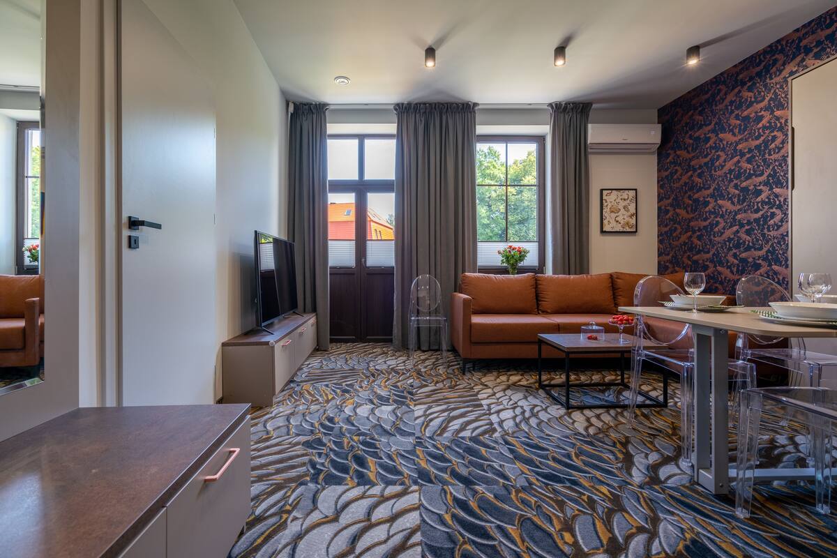 A modern hotel room with a patterned carpet, a sofa, a dining table, and a TV.