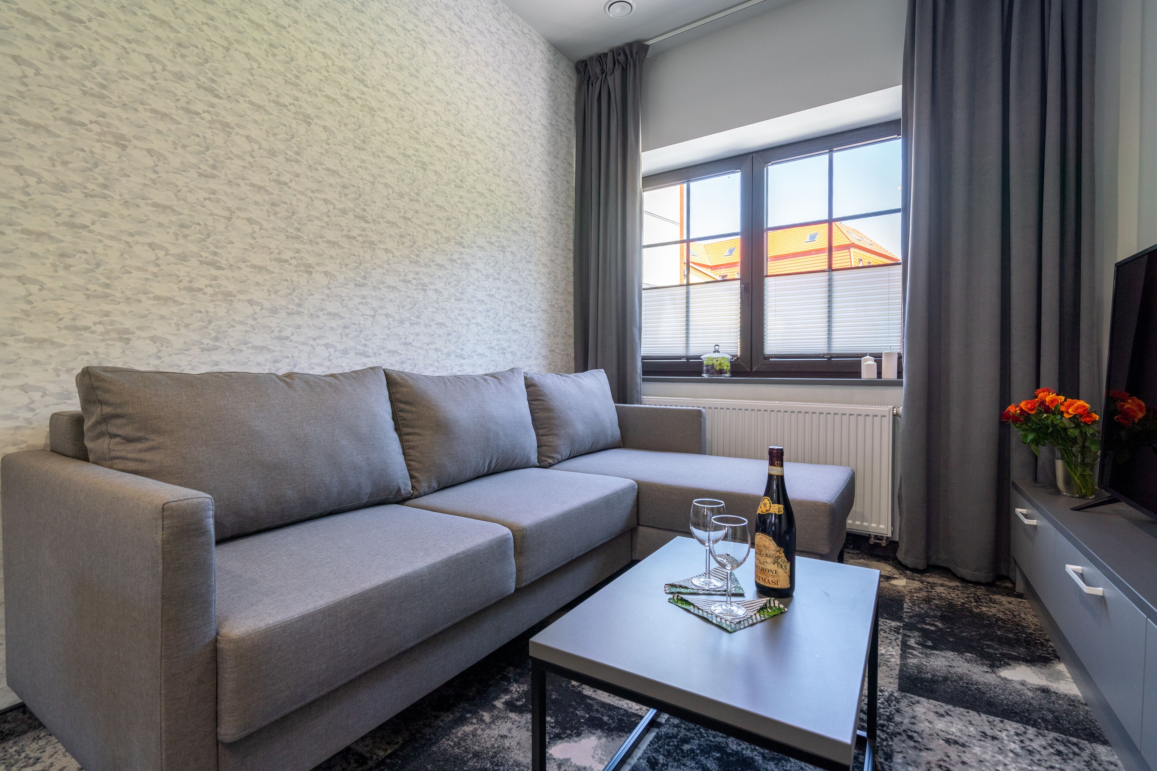 A modern living room with a grey sofa, a small table with a bottle and glasses, a radiator, and a window with curtains.