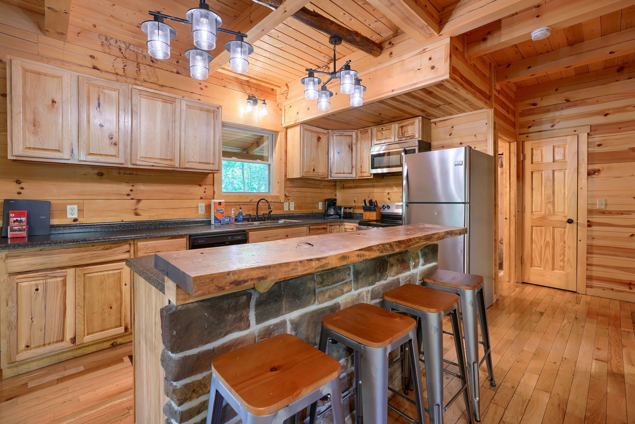 A wooden cabin-style kitchen with a stone accent wall, a stainless steel refrigerator, and a bar area with stools.