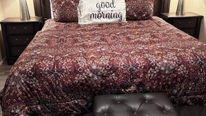 Un lit bien fait, avec une couvre-lit à motifs floraux, un coussin orné de l'inscription « bonjour » et un pouf assorti.