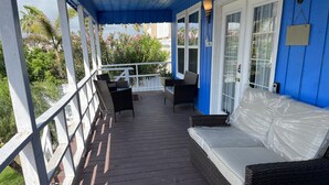 Une véranda couverte avec un plancher en bois foncé, des balustrades blanches et un mur bleu. On y trouve un canapé en osier avec un coussin blanc et une table dont le plateau est de couleur foncée.