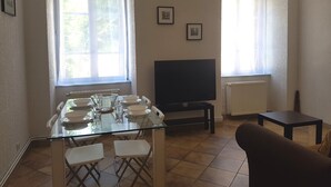 Un coin repas avec une table en verre, des chaises blanches, un téléviseur et un canapé.