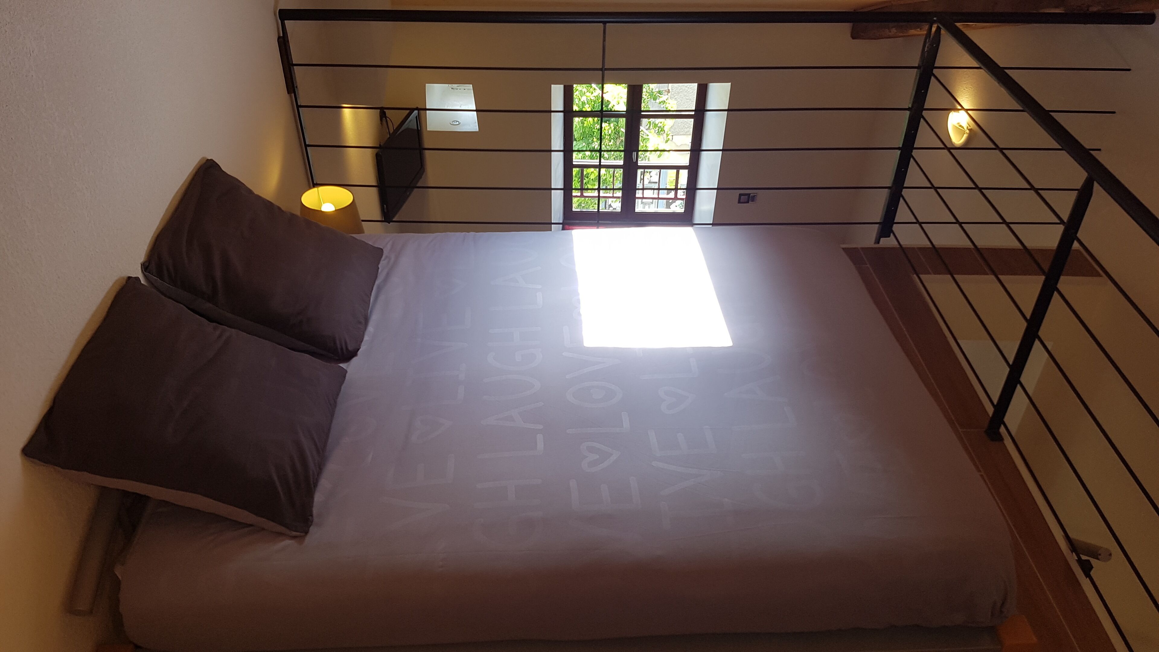 A bed with a light-colored mattress and dark pillows, a window with a view of greenery, and a wall-mounted light fixture.