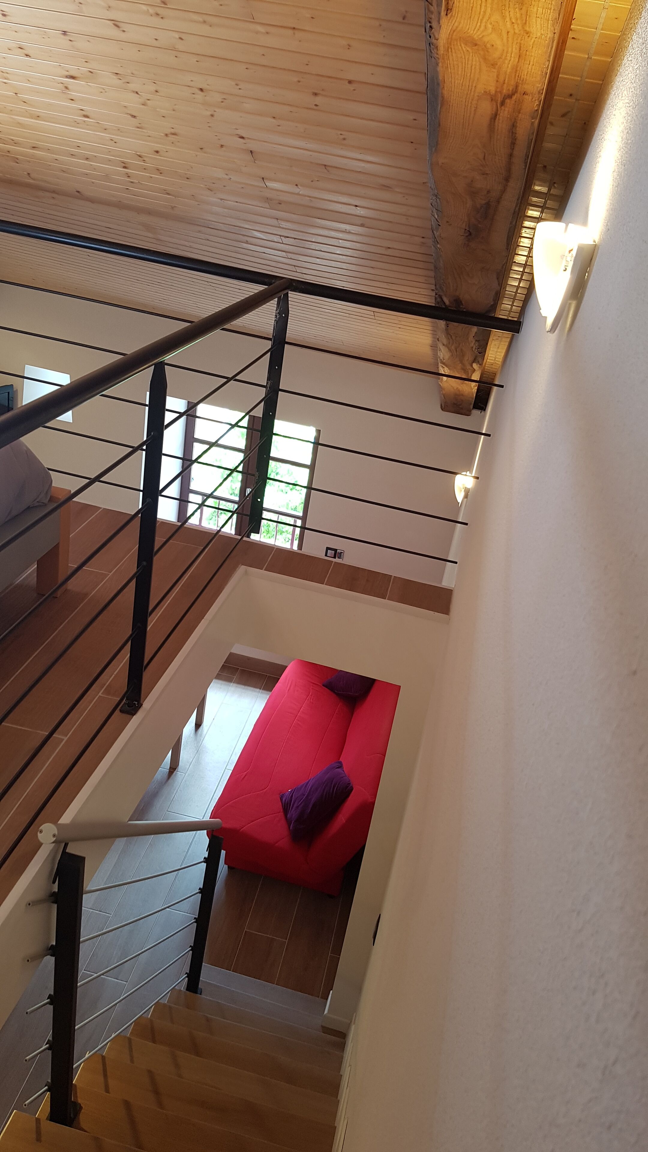 A modern interior with a wooden ceiling, a staircase, a red sofa with a purple pillow, and a white wall.