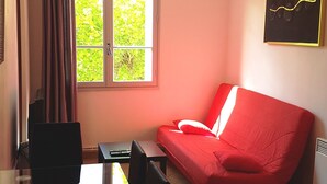 Ein Zimmer mit Esstisch, Stühlen, einem roten Sofa, einem Fenster mit Vorhängen, einer Pendelleuchte und einem gerahmten Bild an der Wand.