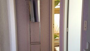 Ein schmaler Flur mit einem Metallregal an der Wand, Fliesenboden und einer Tür zu einem Außenbereich.