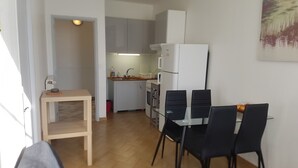 Un ambiente compatto con angolo cottura, una zona pranzo con tavolo in vetro e sedie nere, e un piccolo tavolino con lampada.