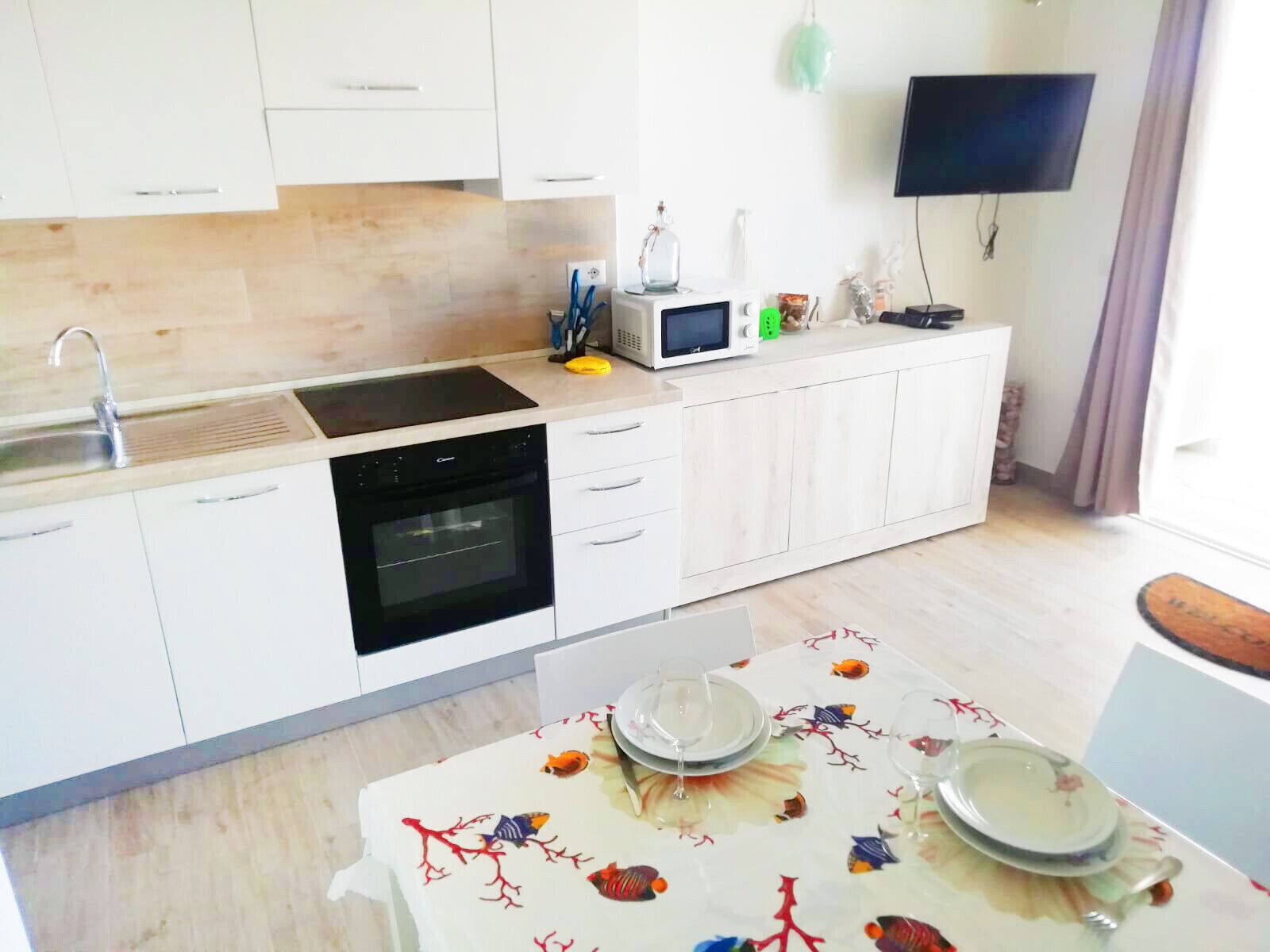A modern kitchen with white cabinets, a built-in oven, a microwave, and a sink. There's a dining area with a table set for two.