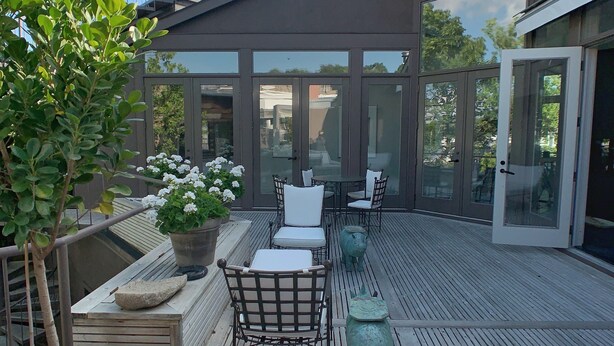 A modern outdoor deck with a glass door leading to a covered area, surrounded by potted plants and outdoor furniture.