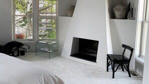 A bedroom with a fireplace, two windows, a bed, a chair, and a small table.