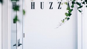 A white door with the name "HUZZA" in black letters, a stone floor, and a white wall with a door on the left.