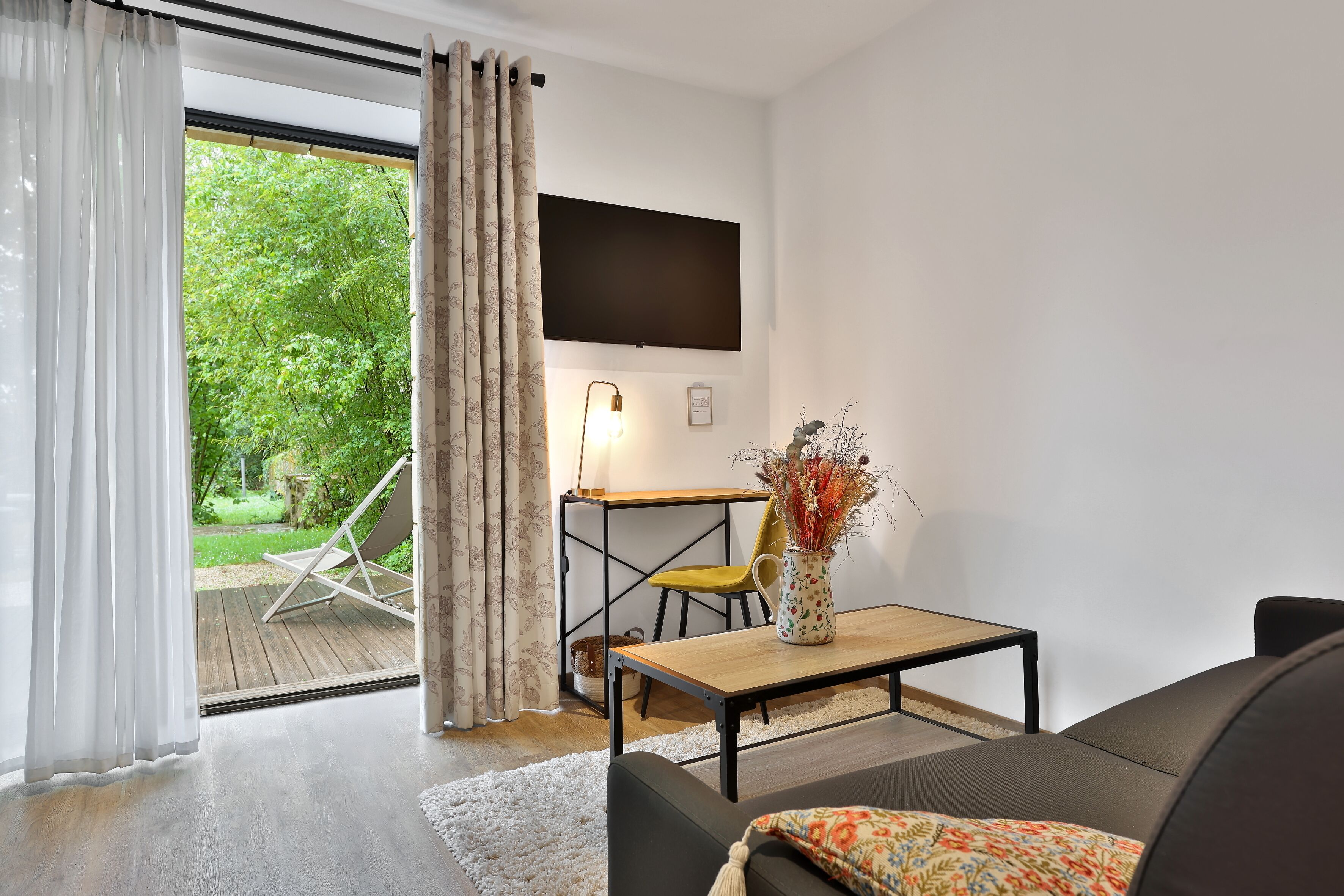 A modern living room with a sofa, a coffee table, and a TV mounted on the wall. There is a window with curtains, a small table with a vase, and a chair.
