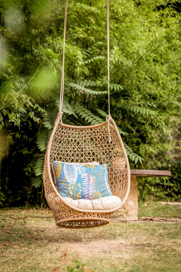 A wicker hanging chair with a cushion, suspended by ropes, in a garden with dense foliage in the background.