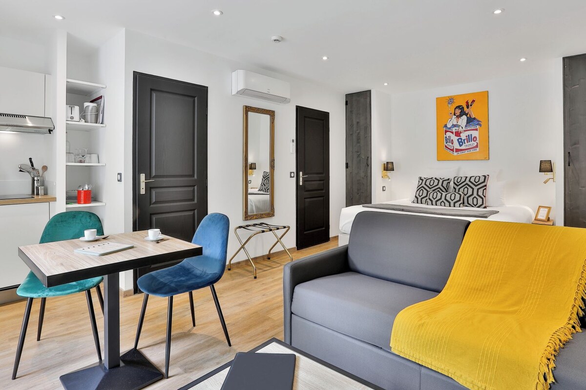Una sala moderna con un sofá gris, una manta amarilla, una zona de comedor con mesa y sillas, y una cocina con una cafetera.