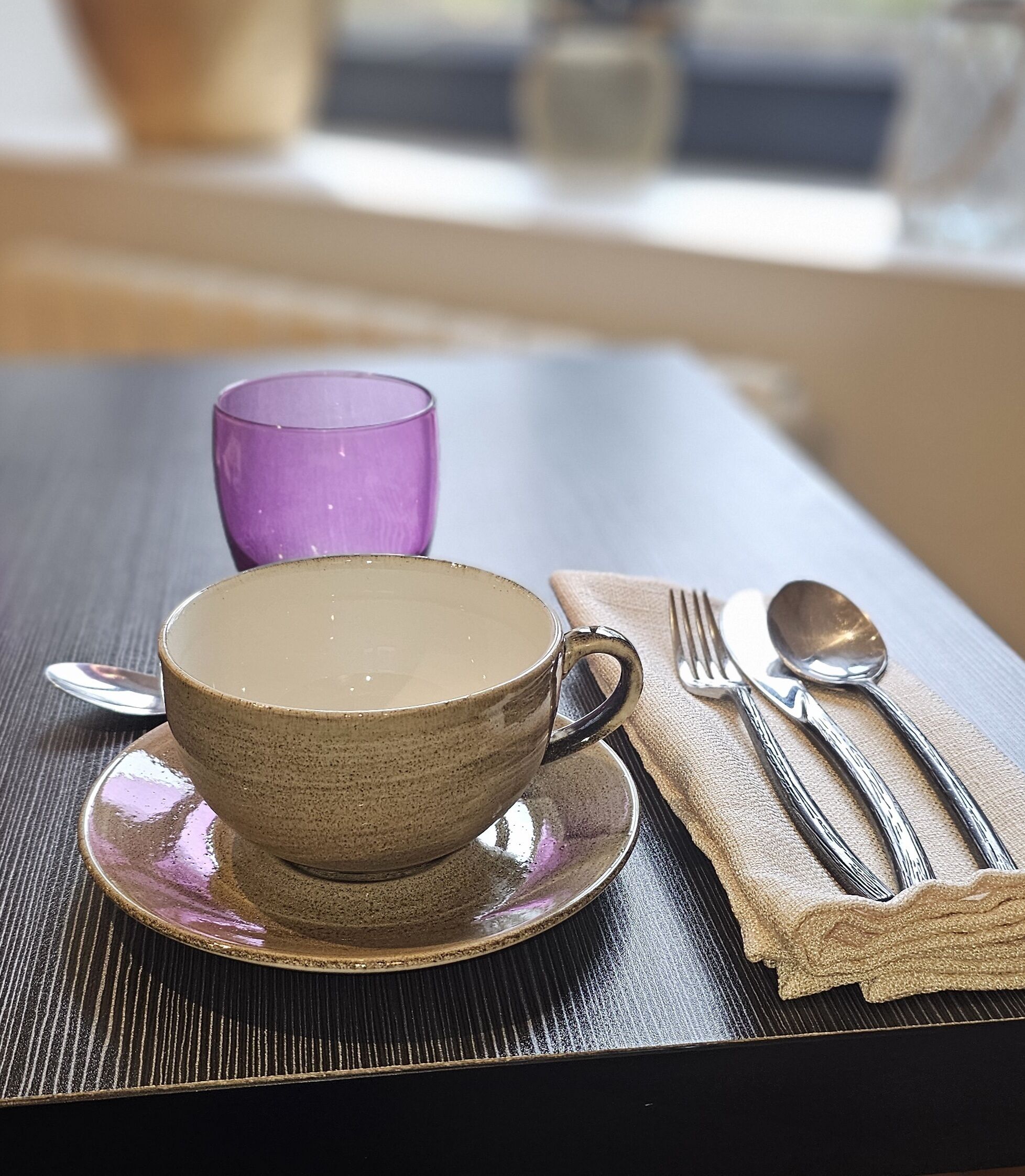 Une table avec une tasse et une soucoupe en céramique, un verre violet et une serviette avec des couverts.