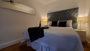 Een net opgemaakt bed met een gestoffeerd hoofdbord, een nachtkastje met een lamp en een dienblad met een fles en glazen.
