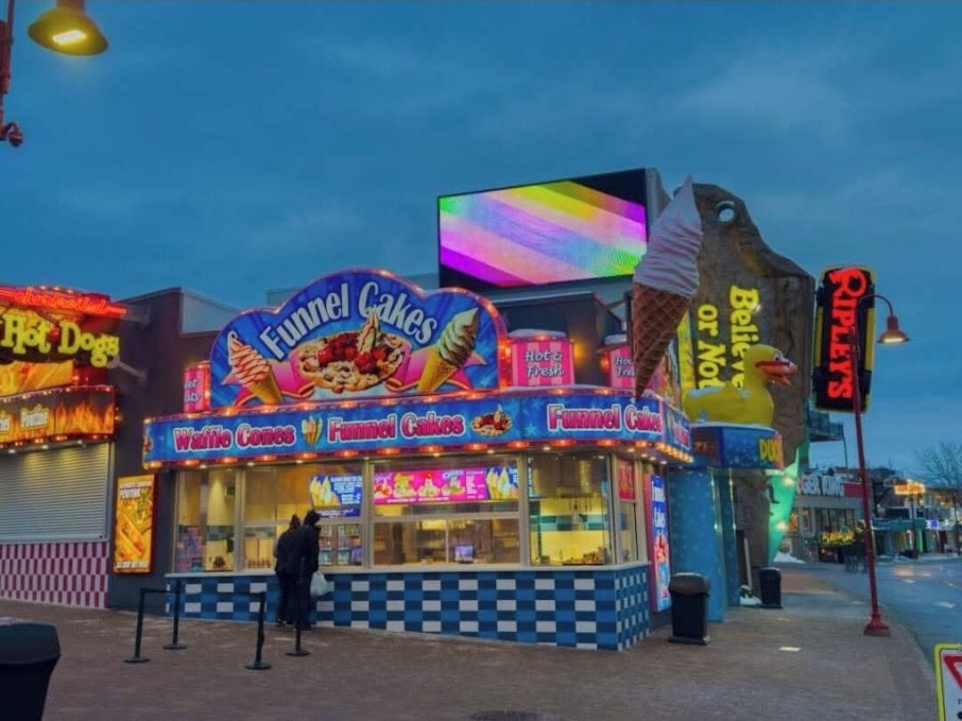 A brightly lit food cart named 'Funnel Cakes' with a neon sign, located on a street corner.