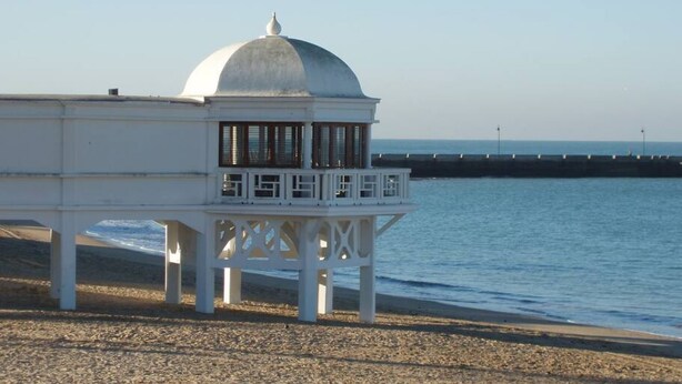 A white pavilion with a dome roof and a balcony, situated on a pebbled beach with a view of the sea and a distant pier. - Apartamento Cadiz de Cine (Cádiz)