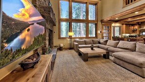 A cozy living room with a fireplace, large windows, a flat-screen TV, and a kitchen area. - Hot Tub, Game Room, Near Lake, Walk to Trails (South Lake Tahoe)