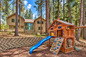 A wooden playhouse with a slide and climbing wall, set against a backdrop of a two-story house and tall pine trees. - Hot Tub, Game Room, Near Heavenly Ski Resort. (South Lake Tahoe)