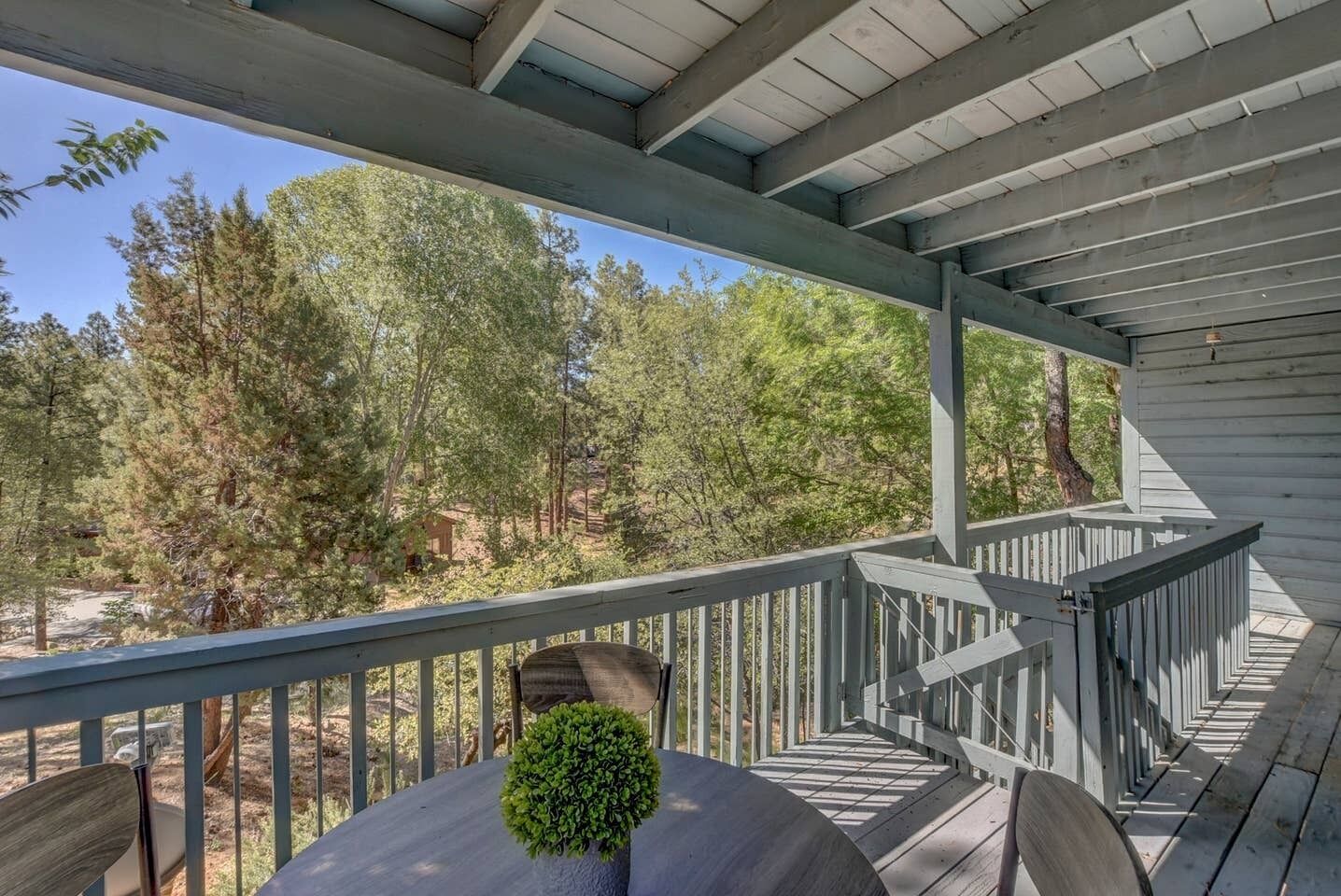 A covered outdoor deck with a railing, a table with a potted plant, and a view of trees and a building in the background.