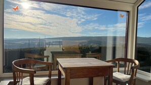 A room with a large window offering a view of a landscape with hills and a body of water. There is a wooden table and two chairs with cushions.