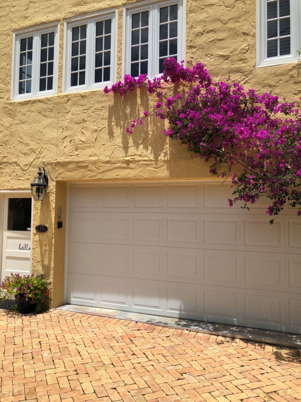 Une maison avec une façade en stuc beige, une porte de garage blanche et des fleurs de bougainvillier violettes.