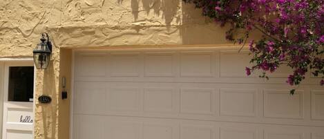 Une maison avec une façade en stuc beige, une porte de garage blanche et des fleurs de bougainvillier violettes.