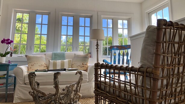 Quarto com sofá branco, cadeira de madeira, ventilador de teto e janelas amplas com vista para o verde.