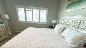 A bedroom with a large bed, a window with blinds, a nightstand with a lamp, and a headboard with a geometric pattern. - Oceanfront townhouse, 2 bedroom, 2.5 baths. (St. Augustine)