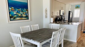 A dining area with a wooden table and white chairs, a kitchen with white cabinets and stainless steel appliances, and a painting of beach umbrellas on the wall. - Oceanfront townhouse, 2 bedroom, 2.5 baths. (St. Augustine)