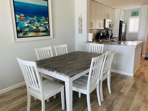 Ein Essbereich mit Holztisch und weißen Stühlen, eine Küche mit weißen Schränken und Edelstahlgeräten sowie ein Bild von Strandsonnenschirmen an der Wand.
