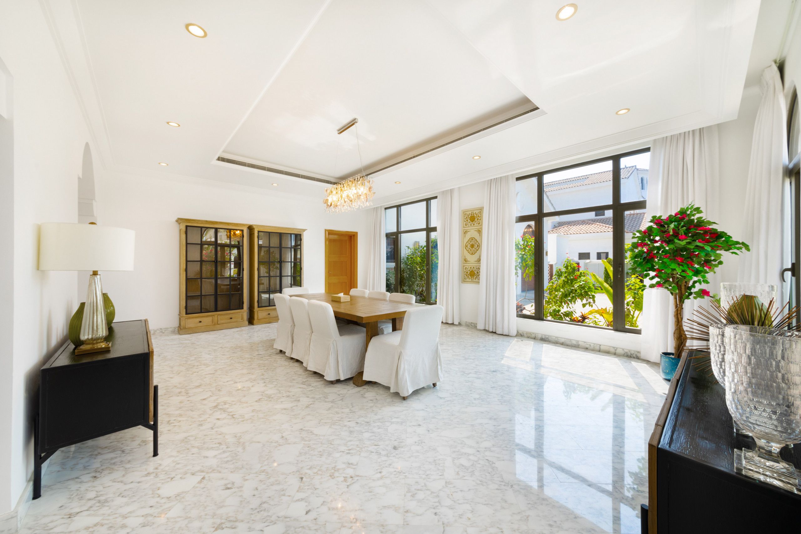 A spacious dining area with a marble floor, a wooden dining table, white chairs, and a chandelier.