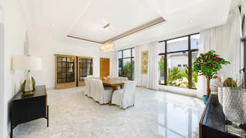 A spacious dining area with a marble floor, a wooden dining table, white chairs, and a chandelier.
