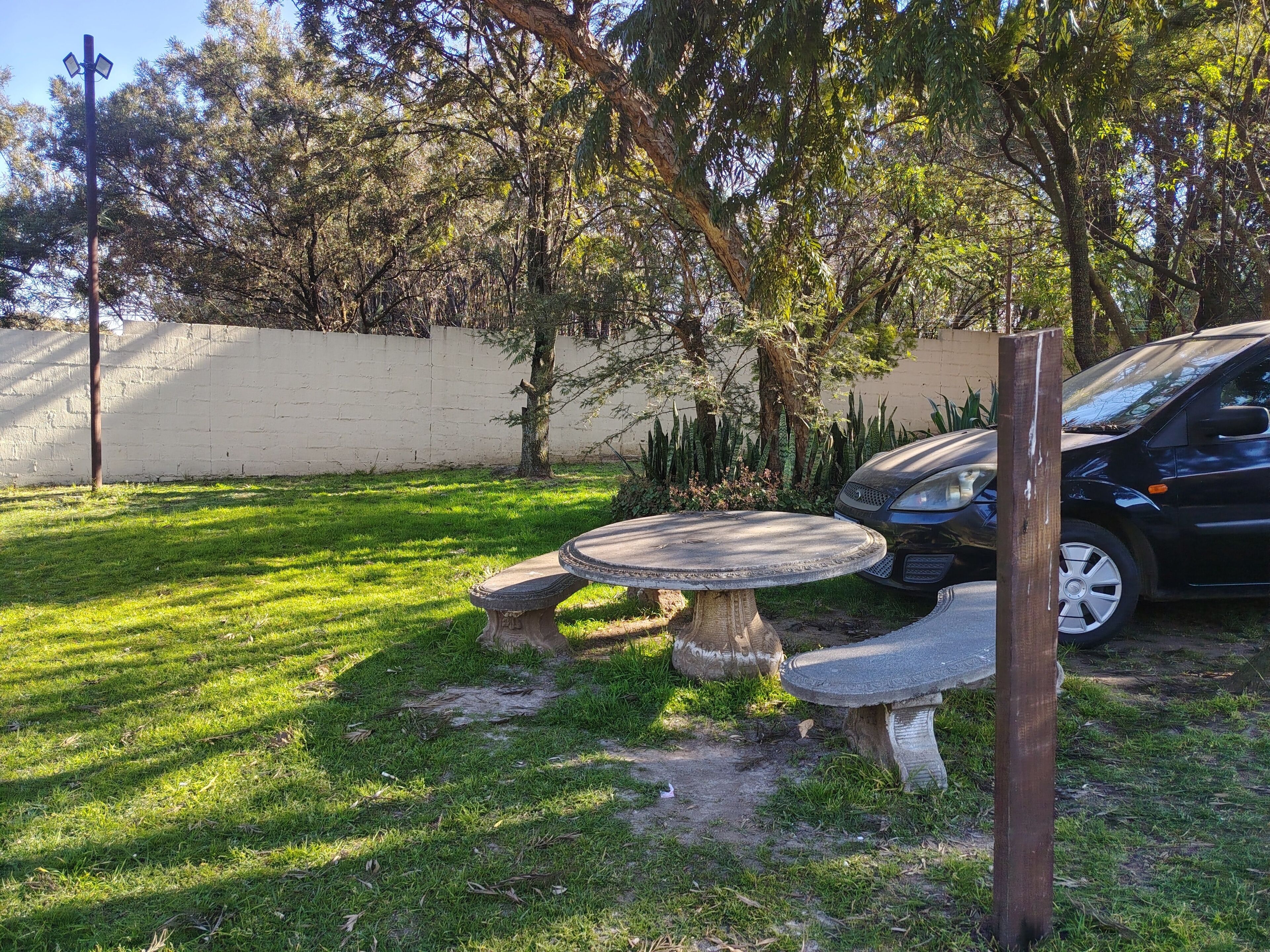 Een tuin met een stenen tafel en banken, omringd door bomen en een geparkeerde auto.