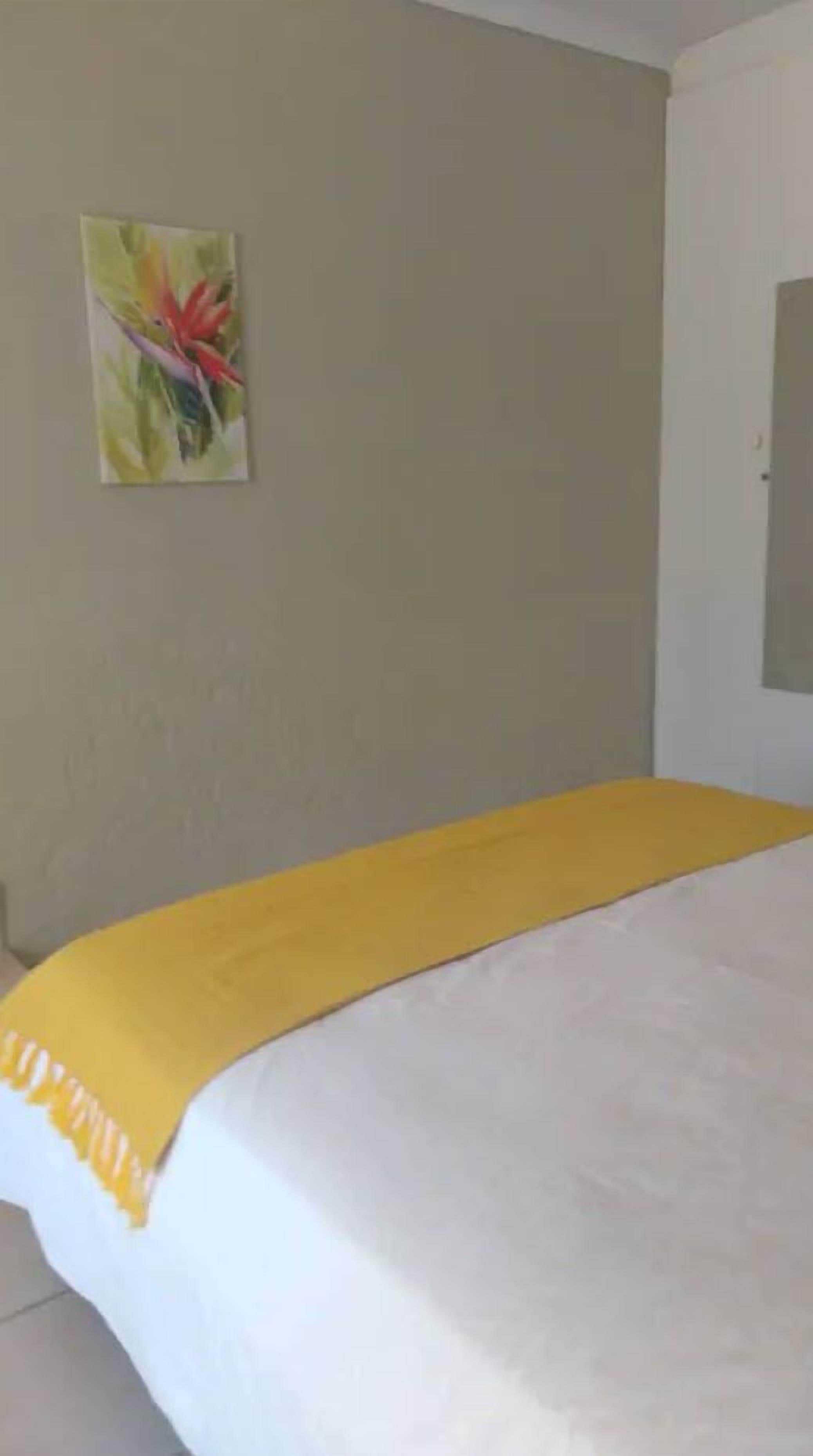 Une chambre d’hôtel avec un lit recouvert d’une couverture blanche et jaune, un tableau accroché au mur et une porte sur le côté droit.